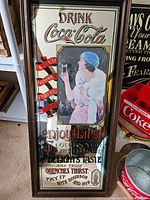 Full frontal view of the mirrored Coca-Cola advertising sign showing the frame and main artwork including the logo, text, and image of the woman.