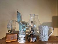 Two hurricane candle holders, a ceramic face pitcher, a blue pheasant statue, a white birdcage, and a wooden box arranged on a shelf.