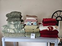 Stack of assorted towels including green patterned, solid green, solid beige, patterned white with red footprint, red, and pinkish towels folded on a table.