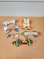 Overview of 11 vintage diecast space toys including Space 1999 Eagle Transporters in white and green, Apollo lunar rover vehicle, and multiple small astronaut and accessory figures, placed on brown surface.