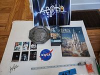 Full lot view showing Apollo 11 Jim Lovell photo promo cards, pewter Alan Shepard plate, Avon Moon Flight game, Apollo 50th Anniversary poster, NASA decal, and booklet.