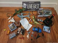 Photo showing an assortment of toys including a James Dean Live Fast metal sign, plastic dinosaur toys, Men in Black toy set, small airplanes, toy vehicles, and various figurines arranged on wood floor.