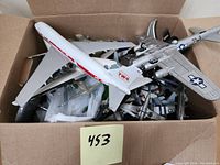 Top view of a cardboard box filled with various unsorted model airplanes including a white TWA commercial plane and a silver military bomber.