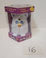 Front and side view of vintage white Furby toy inside original purple and green hexagonal box with plastic window, showing the toy's face and packaging graphics.