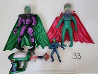 Front view of the three Mars Attacks alien figures showing size differences, detailed sculpting, colored suits, capes, clear helmets, and ray gun toy placed below with ruler for scale.