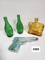 All four uniquely shaped vintage glass bottles seen together: two green Buddha 'Lucky' bottles, one amber whiskey decanter bottle, one clear pistol-shaped bottle with brass cap.