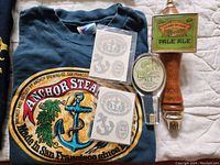 Photo showing vintage dark blue Anchor Steam Beer T-shirt with large logo, two sets of Anchor Steam Beer stickers, Old Foghorn tap handle, and Sierra Nevada Pale Ale tap handle arranged on a white quilt.