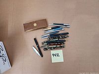 Overall lot on tan background: pen case, modern pens top right, cluster of vintage pens and pencils center