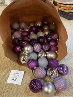 Brown paper bag filled with approximately 37 plastic Christmas ball ornaments in purple, lavender, and silver color tones. Ornaments vary in finish from shiny metallic, matte, ribbed texture, to glitter-coated. Each approximately 3 inches in diameter.