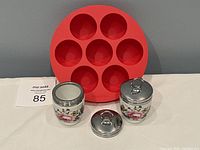 Three Royal Worcester porcelain egg coddlers with floral designs and metal lids, alongside a red silicone Instant Pot egg cooker with seven compartments.
