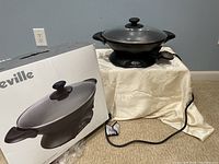 Breville electric wok with glass lid and black handles shown with original box. Temperature dial and power cord visible, unit placed on a beige cloth.