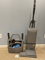 Front view of Electrolux upright vacuum cleaner next to a plastic storage bin holding vacuum attachments including hose and brushes.