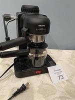 Full view of the black Krups espresso maker with glass carafe and power cord, sitting on table with lot card number 73.