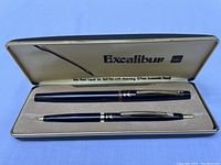 Open box view showing the black Excalibur liquid ink ball pen and 0.7mm mechanical pencil resting inside the beige velvet-lined interior.