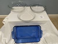 Four glass baking dishes displayed on a white cloth: one blue rectangular baking dish front center, two clear glass pie pans on the right, one larger clear glass bowl on the left