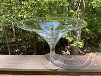 Clear glass display bowl shown outdoors with trees in background, showcasing thick glass and wide flared rim.