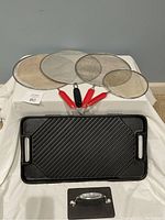 Four mesh splatter screens of varying sizes with red and black handles and a reversible cast iron grill/griddle on a white cloth.