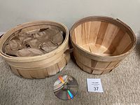 Two wooden harvest baskets side by side on carpeted floor, round basket filled with brown paper and oval basket empty; a CD placed in front for size reference.
