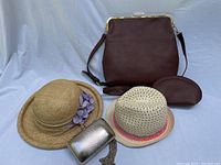 Full lot showing four hats/purses: large brown vegan handbag with shoulder strap, child's woven straw hat with pink trim, adult beige straw hat with purple flower decor, and gold metallic evening clutch. Also small brown zippered pouch is present.