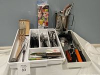 Wide photo showing assortment of cutlery, sharpener, straws, and various kitchen utensils in a white tray with utensil holder in background.