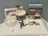 Kitchenmate manual food processor components including bowl, lid with hand crank, white plastic basket, three blade attachments, original box, and instruction manual laid out on table.