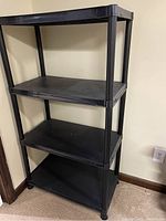 4-tier black plastic shelf unit against beige wall, showing four shelves and cylindrical legs.