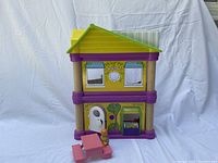 Front view of Play Town two-story doll house with yellow roof, purple accents, white door, and clock detail, plus pink table and chair and yellow figurine in front.