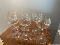 Six large red wine glasses placed on a wooden surface, showing their broad bowl shape and clear glass design.