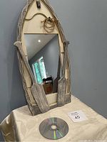 Front view of the rowboat-shaped mirror standing upright on a draped surface with a CD placed next to it for size reference. The mirror has wooden oars attached on both sides and rope detail at the top.