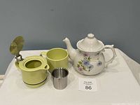 Porcelain tea set including white floral teapot and green single serve brewing cup with lid open and stainless steel strainer placed beside it.