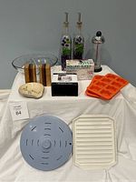 Full view of lot items arranged on white draped surface showing multiple kitchen utensils and containers including glass dispensers, wooden shakers, trays, and plastic covers.