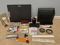 Various office supplies including bean bag lap desk, file organizer, pens, pencils, stapler, rulers, tape dispenser, and other desk accessories arranged on carpeted floor