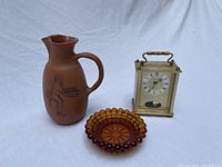 Three items shown together: brass carriage clock, amber glass candle holder, terracotta pitcher with centaur illustration.