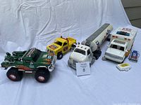 Four toy trucks arranged left to right: green monster truck, yellow Chevy pickup truck, Chevron gasoline tanker truck, white Hess rescue ambulance truck with accessories in front.