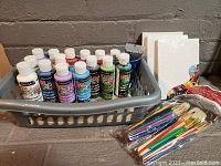 Photo showing multiple bottles of DecoArt Crafter's Acrylic Paint in a gray basket next to wrapped canvases and package of brushes
