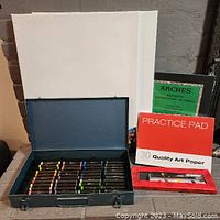 Photo showing blue metal case of pastels in front of two white canvases and two art paper pads.