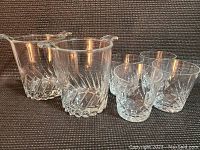 Photo showing four clear glass whiskey tumblers and two glass ice buckets on fabric background, highlighting their clear glass and textured designs.