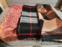 Three scarves laid flat showing varied patterns and colors: red paisley, black/grey/red plaid cashmere, and brown geometric.