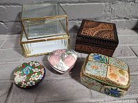 Collection of five trinket boxes displayed together including glass and brass box, black lacquered box, heart-shaped pink floral box, colorful round cloisonné box, and hand-painted square box.