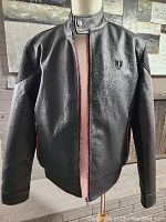 Front view of black faux leather men's jacket showing stand collar with snap button and red lining visible on zipper lips.