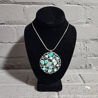 Front view of necklace displayed on black stand showing round wire-wrapped pendant with multiple polished stones including turquoise.