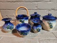 Six-piece vintage ceramic breakfast set arranged on a floor. Includes creamer, sugar bowl, jam pot, and small vases, all with blue and white glaze and raised berry and leaf detail.