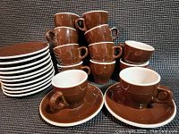 Set of six brown porcelain espresso cups arranged in two stacks alongside six matching saucers stacked separately. Cups have a glossy finish and white interior rims.