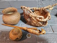 Lot with two woven Indigenous baskets, one Haida carved letter opener, and one hand-etched dyed gourd placed on a stone surface with coin for size reference.