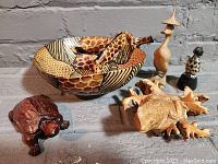 Six different wildlife-themed wood carvings arranged on a surface: a large bowl with giraffe pattern, two carved crocodiles, a dark reddish-brown turtle, and two upright bird figures, one black and white and one light-colored with a hat.