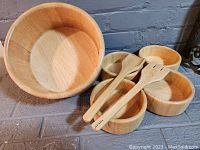 Set of 1 large bamboo salad bowl, 4 smaller serving bowls and 2 wooden salad utensils placed on floor against brick wall.