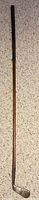 Full image of antique wooden shafted golf club placed on carpeted floor, showing entire length including wooden shaft and metal blade.