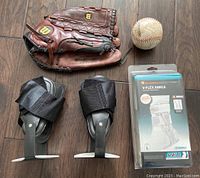 Photo of glove, softball, used and unopened ankle braces laid flat on wooden surface.