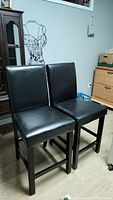 Two black leather-like upholstered bar chairs showing front view, a pair, solid shape with some scratches.