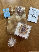 Photo showing two items: clear crystal sun catcher pendant with hanging wire and suction cup, gold satin pouch, and decorative flower-shaped brooch with purple stones in box.